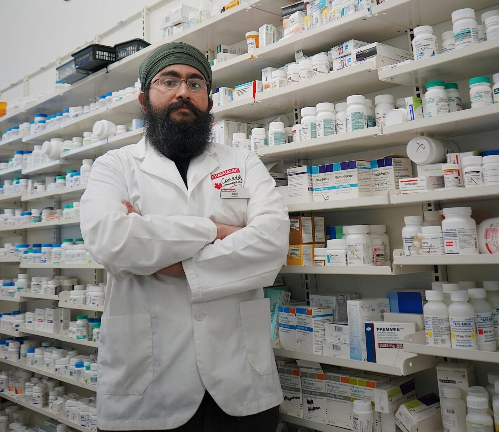 Pharmacist with turban and glasses in a pharmacy, standing in front of medicine shelves.